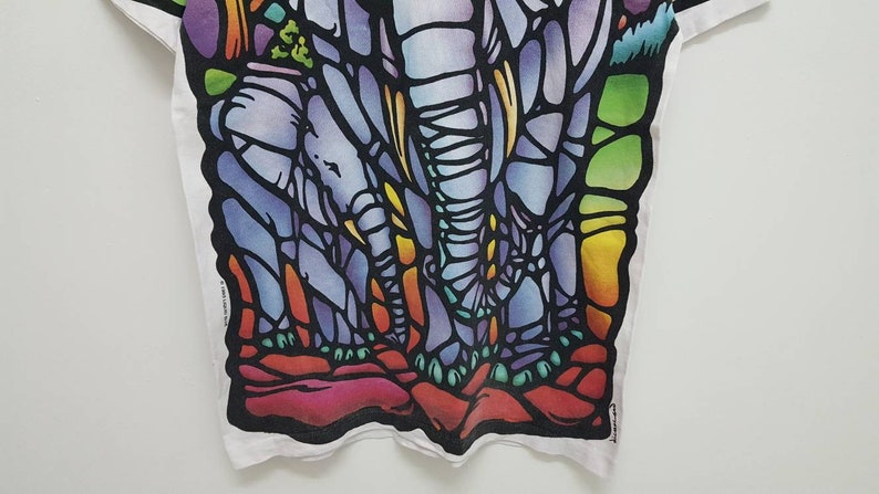 Puede incluir: Una camiseta blanca con un dise&ntilde;o de vitral colorido de tres elefantes. Los elefantes est&aacute;n delineados en negro y rellenos con tonos de azul, morado, verde, amarillo y naranja.