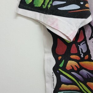 Puede incluir: Camiseta blanca con un estampado gr&aacute;fico abstracto de colores. El dise&ntilde;o presenta una variedad de formas y colores, incluyendo rojo, naranja, amarillo, verde, azul y morado. El gr&aacute;fico parece ser una representaci&oacute;n estilizada de una persona o un grupo de personas.