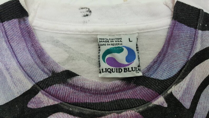Puede incluir: Camiseta blanca con estampado abstracto morado, negro y blanco. La camiseta tiene una etiqueta de Liquid Blue con el texto "100% algod&oacute;n hecho en EE. UU. cuidado en el reverso" y una talla L.