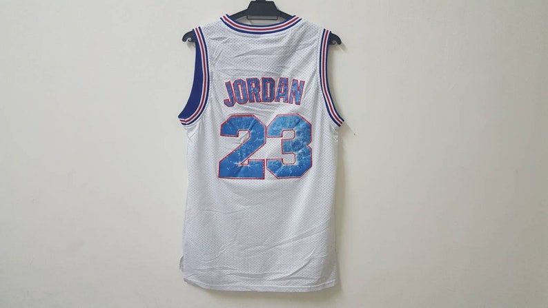 May include: White basketball jersey with blue and red trim. The back of the jersey has the name "JORDAN" and the number "23" in blue with a white outline.