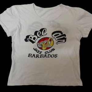 May include: White t-shirt with a round neck and short sleeves. The shirt features a graphic with the words "Bee on" in black script above a red and yellow jazz instrument design. Below the design, the text "Jazz Club Barbados" is printed in black.