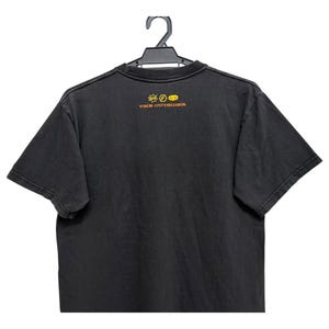 Puede incluir: Camiseta negra colgada de una percha negra. La camiseta tiene un aspecto descolorido y presenta un peque&ntilde;o gr&aacute;fico y el texto "THE OUTSIDER" en letras doradas en la parte superior de la espalda. La camiseta es de manga corta.