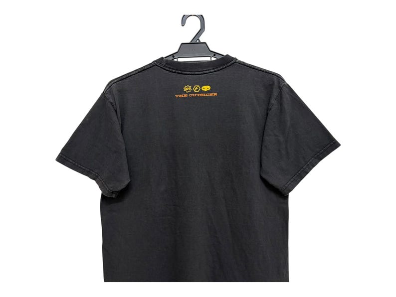 Puede incluir: Camiseta negra colgada de una percha negra. La camiseta tiene un aspecto descolorido y presenta un peque&ntilde;o gr&aacute;fico y el texto "THE OUTSIDER" en letras doradas en la parte superior de la espalda. La camiseta es de manga corta.