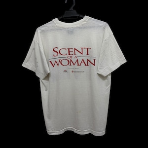 May include: A white short-sleeved t-shirt featuring the red text "SCENT OF A WOMAN" printed on the back. Below the title, there are two small logos, one of which includes the text "BAKER & TAYLOR" in red, displayed on a plain background.