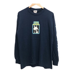 May include: A navy blue long-sleeved t-shirt featuring a graphic of a person in a blue and yellow box. The text "Stella" is printed in yellow above the box.