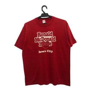 May include: Red t-shirt with the text "Ronald McDonald House Iowa City" in white lettering. The design features a heart shape. The shirt is hanging on a black hanger.