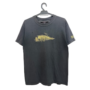 Könnte beinhalten: Ein anthrazitfarbenes Kurzarm-T-Shirt mit einem goldfarbenen Grafikdruck auf der Brust, der in stilisierter Schrift Atticus liest. Der gleiche goldfarbene Schriftzug befindet sich auch auf dem linken Ärmel. Das T-Shirt hängt an einem schwarzen Kleiderbügel.