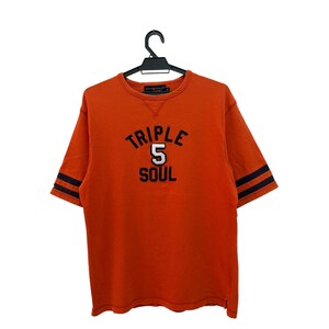 Könnte beinhalten: Orangefarbenes T-Shirt mit kurzen Ärmeln und dem Text "TRIPLE 5 SOUL" in Marineblau und Weiß. Die Ärmel haben marineblaue Streifen. Das T-Shirt hängt an einem schwarzen Kleiderbügel.