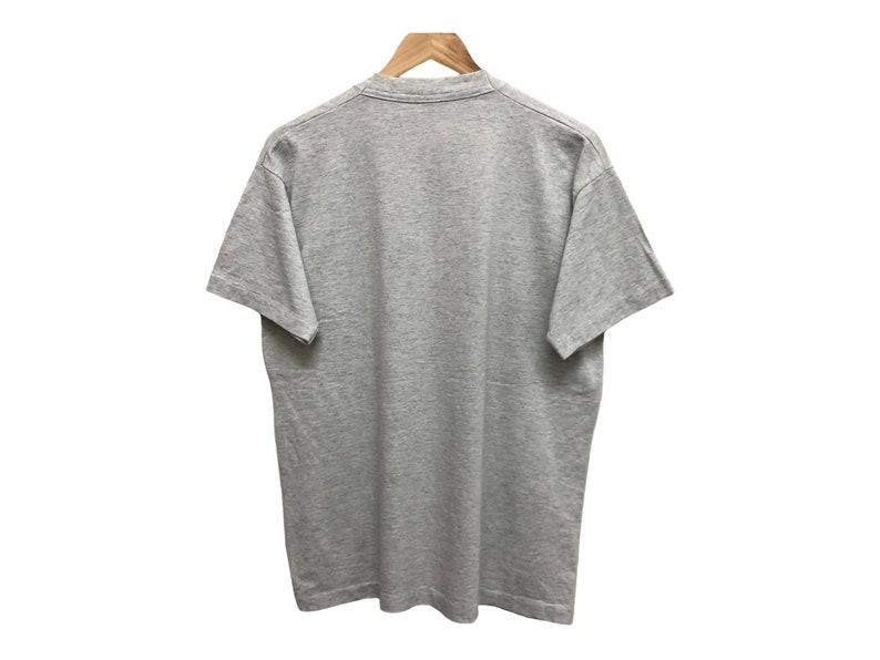 Op de afbeelding: Een grijs t-shirt met korte mouwen. Het t-shirt hangt aan een houten hanger.
