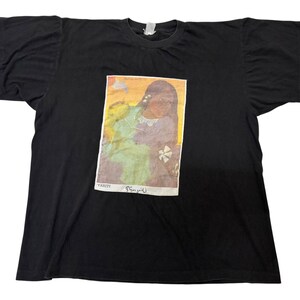May include: A black short-sleeve t-shirt featuring a rectangular graphic print of a painting depicting a person in a light green garment. The print includes the text TAHITI and the signature P. Gauguin at the bottom of the rectangular image.