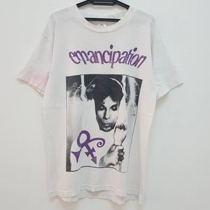 May include: White t-shirt with a black and white photo of Prince on the front. The photo is surrounded by a purple border and the word "emancipation" is printed in purple above the photo.