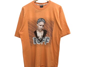 Op de afbeelding: Oranje T-shirt met een grafische print van een vrouw met een geschoren hoofd en het woord "RUFF" eronder gedrukt. Het shirt heeft een ronde hals en korte mouwen.