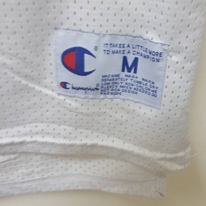 May include: White Champion brand mesh jersey with a blue and white tag that reads "It takes a little more to make a champion" and "M" for size. The tag also includes care instructions.