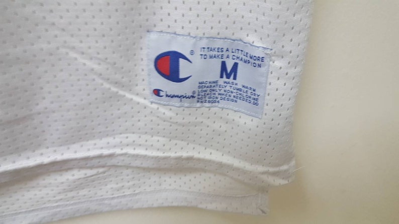 May include: White Champion brand mesh jersey with a blue and white tag that reads "It takes a little more to make a champion" and "M" for size. The tag also includes care instructions.