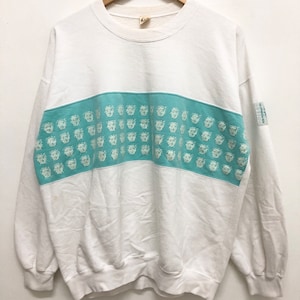 Puede incluir: Sudadera blanca con una banda azul turquesa a través del pecho. La banda tiene un patrón repetitivo de caras blancas.