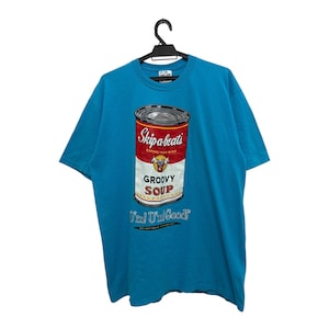 以下が含まれることがあります： ターコイズ色のTシャツ。スープ缶のグラフィックが描かれており、赤、白、黒で「Skip a beats GROOVY SOUP」と書かれています。シャツの下部には「Feel U're Good!」の文字も入っています。