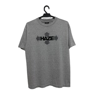 Puede incluir: Camiseta gris con la palabra "HAZE" en negro y flechas que apuntan en cuatro direcciones. La camiseta está colgada de una percha negra.