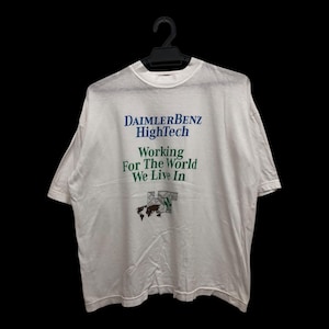 Op de afbeelding: Wit T-shirt met de tekst "DAIMLERBENZ HighTech" in blauw en "Working For The World We Live In" in groen. Er is ook een wereldkaartafbeelding aanwezig. Het shirt hangt aan een zwarte hanger.