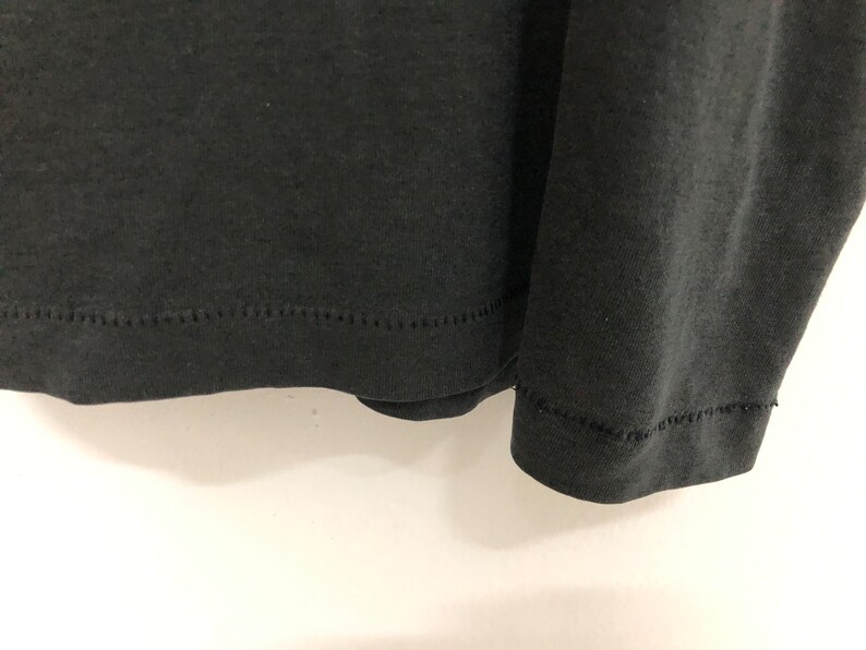 May include: Close-up of the bottom hem of a black long-sleeved shirt. The hem is stitched with a zig-zag stitch.