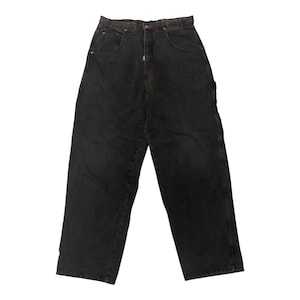 Puede incluir: Un par de vaqueros negros. Los vaqueros tienen un diseño clásico de cinco bolsillos, cierre de botón y cremallera, y un corte relajado. El tejido vaquero parece ligeramente descolorido, lo que le da un aspecto desgastado. Los vaqueros se muestran sobre un fondo blanco.