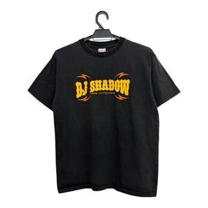 Puede incluir: Camiseta negra con el texto "BJ SHADOW" en amarillo y naranja, con "THE OUTSIDER" debajo. La camiseta est&aacute; colgada de una percha negra.