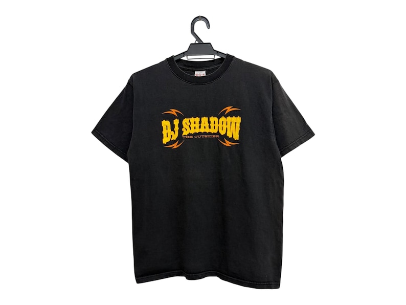 Puede incluir: Camiseta negra con el texto "BJ SHADOW" en amarillo y naranja, con "THE OUTSIDER" debajo. La camiseta est&aacute; colgada de una percha negra.