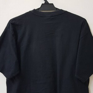 K&ouml;nnte beinhalten: Schwarzes T-Shirt mit kurzen &Auml;rmeln, das an einem schwarzen Kleiderb&uuml;gel h&auml;ngt. Der &Auml;rmel tr&auml;gt das Wort "PRAGUE" in wei&szlig;en, blauen und roten Buchstaben. Das Shirt ist aus einem weichen, dunklen Stoff gefertigt.