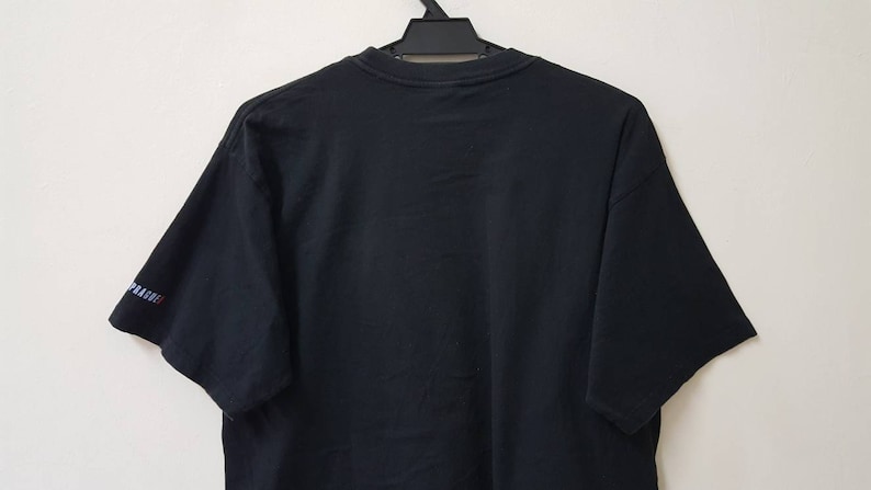 K&ouml;nnte beinhalten: Schwarzes T-Shirt mit kurzen &Auml;rmeln, das an einem schwarzen Kleiderb&uuml;gel h&auml;ngt. Der &Auml;rmel tr&auml;gt das Wort "PRAGUE" in wei&szlig;en, blauen und roten Buchstaben. Das Shirt ist aus einem weichen, dunklen Stoff gefertigt.