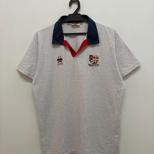 May include: A light grey polo shirt with a navy blue collar and red trim. The shirt has embroidered logos on the chest, including the word "HASBRO" and a crest. The short-sleeved shirt is hanging on a black hanger.