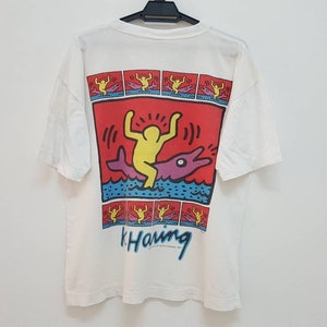 K&ouml;nnte beinhalten: Wei&szlig;es T-Shirt mit einem bunten Grafikdruck einer Person, die einen Delfin reitet. Die Grafik ist von einem roten Rand umgeben und enth&auml;lt den Text "k. Haring".