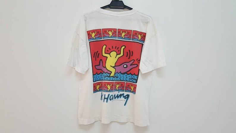 K&ouml;nnte beinhalten: Wei&szlig;es T-Shirt mit einem bunten Grafikdruck einer Person, die einen Delfin reitet. Die Grafik ist von einem roten Rand umgeben und enth&auml;lt den Text "k. Haring".