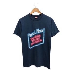 May include: A dark blue t-shirt with a white and blue neon sign graphic that says "Pepsi Now! The choice of a new generation."
