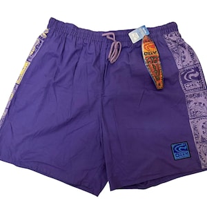 May include: Purple swim trunks with a drawstring waist and decorative patterned panels on the sides. The panels feature a repeating design in shades of purple, yellow, and white. A tag with a surfboard design is attached.