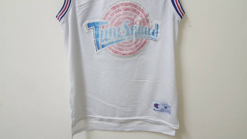 May include: White basketball jersey with a blue, red, and white "Tune Squad" logo on the front. The jersey is made of mesh fabric and has a Champion logo on the bottom right.