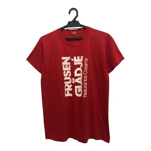 Op de afbeelding: Rood t-shirt met de tekst "FRUSEN GLÄDJÉ Natural Ice Creams" in het wit. Het shirt hangt aan een zwarte hanger. De tekst is verticaal uitgelijnd op de voorkant van het shirt.