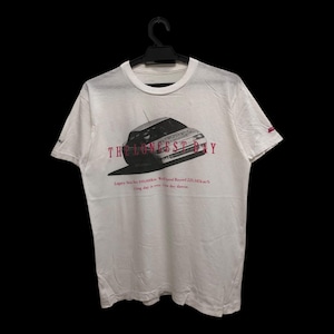 May include: White t-shirt with a graphic of a car and the text "THE LONGEST DAY" in maroon. Additional text below the car graphic. The shirt is hanging on a black hanger.