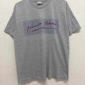 May include: A gray t-shirt with the text "Pointer Sisters" in a red script font, set within a blue grid. The shirt is hanging on a black hanger. The shirt is made of a soft material.