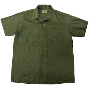 May include: Olive green short-sleeved shirt with a pointed collar and two chest pockets. The shirt has a button-down front and a small tag near the collar. The fabric has a subtle textured pattern.