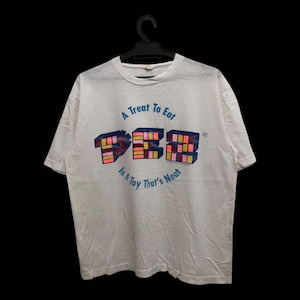 May include: White t-shirt with a colorful graphic of the PEZ logo and the text "A Treat To Eat In A Toy That's Neat". The graphic features the PEZ logo in blue, pink, yellow, and orange.
