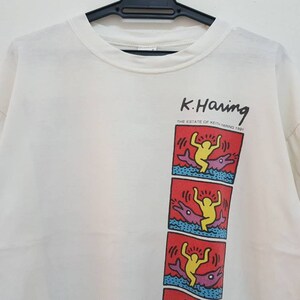 K&ouml;nnte beinhalten: Wei&szlig;es T-Shirt mit einem Grafikdruck eines Cartoon-Charakters, der auf einem Delfin reitet. Der Text "K. Haring" ist &uuml;ber dem Grafikdruck gedruckt. Der Text "THE ESTATE OF KEITH HARING 1989" ist unter dem Grafikdruck gedruckt.