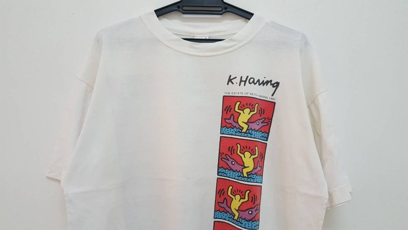 K&ouml;nnte beinhalten: Wei&szlig;es T-Shirt mit einem Grafikdruck eines Cartoon-Charakters, der auf einem Delfin reitet. Der Text "K. Haring" ist &uuml;ber dem Grafikdruck gedruckt. Der Text "THE ESTATE OF KEITH HARING 1989" ist unter dem Grafikdruck gedruckt.