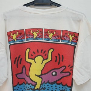 K&ouml;nnte beinhalten: Wei&szlig;es T-Shirt mit einem bunten Grafikdruck einer Person, die auf einem Delfin reitet. Die Grafik ist von einem roten Rand umgeben und enth&auml;lt den Text "!!!".