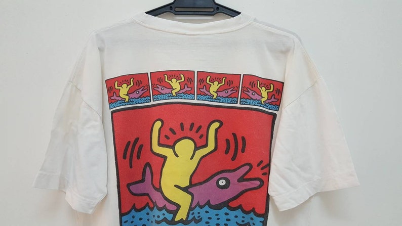 K&ouml;nnte beinhalten: Wei&szlig;es T-Shirt mit einem bunten Grafikdruck einer Person, die auf einem Delfin reitet. Die Grafik ist von einem roten Rand umgeben und enth&auml;lt den Text "!!!".