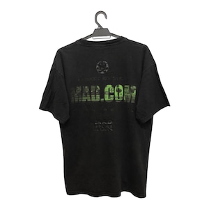 Op de afbeelding: Zwart t-shirt met de groene tekst "MAD.COM" en andere vervaagde afbeeldingen. Het shirt hangt aan een zwarte hanger tegen een witte achtergrond.