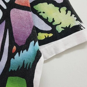 Puede incluir: Una camiseta negra con un estampado abstracto de colores. El estampado presenta una variedad de formas y colores, incluyendo morado, azul, verde y amarillo. La camiseta tiene un ribete blanco alrededor del cuello y las mangas.