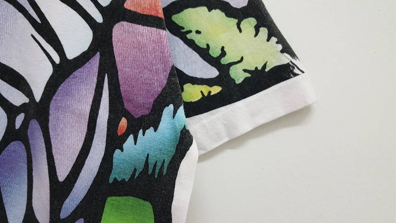 Puede incluir: Una camiseta negra con un estampado abstracto de colores. El estampado presenta una variedad de formas y colores, incluyendo morado, azul, verde y amarillo. La camiseta tiene un ribete blanco alrededor del cuello y las mangas.