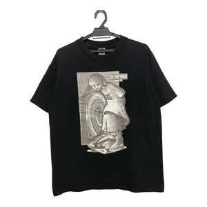 May include: Black t-shirt featuring a graphic print of a person wearing a white shirt and black trousers, standing in front of a large circular object. The text "DAY ONE" is printed on the graphic.