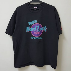 Könnte beinhalten: Schwarzes T-Shirt mit einem Aufdruck von "Tom's Hard Luck Cafe Henderson" in einem Farbschema aus Lila, Blau und Rosa.