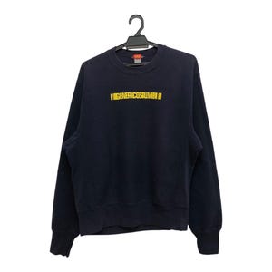 May include: A navy blue sweatshirt with the yellow text "IGENERICOSIUMBI". The long-sleeved top is suspended from a black hanger against a white backdrop. The sweatshirt features a crew neck and ribbed cuffs and hem.