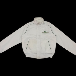 Op de afbeelding: Witte jas met een opstaande kraag en een groen logo op de borst. De jas heeft een ritssluiting, twee voorzakken en groene biezen langs de naden. De mouwen zijn lang en de zoom is elastisch.